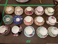 Top-down view of all 14 assorted fine bone china teacups and saucers laid out on a table, showing variety of colors and patterns.