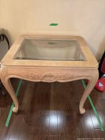 Front-facing photo of light wood tone side table with square glass top. Shows carved shell design on apron and cabriole legs.