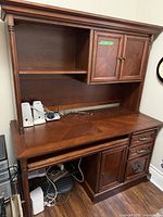 Front view of the wooden office desk showing the work surface, hutch with cabinet doors and open shelf, and electronic cables underneath.
