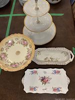 Three-tiered serving stand and three ceramic serving dishes with floral patterns and ornate edges on dark surface.