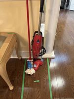 Front view of red Vileda steam mop standing upright with reusable white mop cloth attached to triangular mop head. Mop is shown against a hardwood floor and next to a table and pillar.