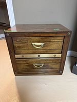 Front view of small wood dresser showing two drawers with gold handles and decorative metal strip.