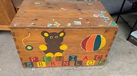 Top and front view of wooden toy chest with painted bear, colorful ball, letter blocks, and stickers on surface.