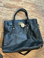 Front view of the Michael Kors black leather handbag placed on a wooden surface, showing the gold-tone lock and hanging tag on front.