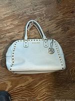 Front view of white leather Michael Kors satchel on wood surface, showing gold studs, logo, and charm