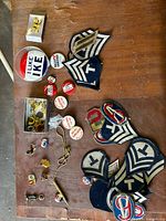 Top view of collection on wooden surface showing large 'I Like Ike' button, numbered military patches, smaller pins, and pin storage box with parts.