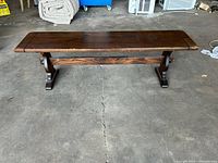 Top front view of a long wooden bench showing dark brown stained wood and trestle-style legs on a concrete floor.