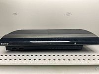 Front view of black Sony PlayStation 3 gaming console on perforated metal shelf.