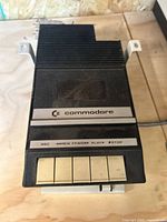 Top-down view of black Commodore cassette drive, branded with logo, showing push-button controls and mounting brackets.