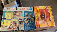 Lot showing two vintage Easy-Bake Oven boxes, one yellow and one orange, with children's images and cookie baking graphics on the packaging.
