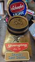 Photo showing collection of vintage bar memorabilia including a round Schaefer Light sign, embossed Löwenbräu barrel top, and multiple Yuengling signs laid on a wooden surface.