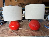 Two West Elm table lamps with glossy red ceramic bases and beige fabric shades placed side by side on a wooden surface.