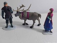 Photo showing Anna, Kristoff, and Sven vinyl figurines on a white surface with visible snow bases, displaying the front and side profiles of the characters.