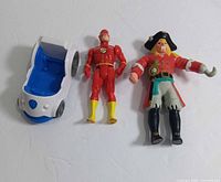 Photo showing all three items in the lot: The Flash action figure in red, Pirates of the 7 Seas pirate figure with hook, and the small Paw Patrol blue and white vehicle.
