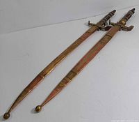 Pair of brass and copper decorative swords with wood and steel handles, sheathed in decorated scabbards, laid side by side.