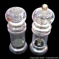 Left to right view showing the salt shaker with multi-hole top and pepper mill with metallic components inside and on top, both clear acrylic, standing upright on black background.