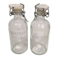 Pair of vintage clear glass toilet water bottles with white ceramic stoppers and wire clips shown side by side. Both bottles have white printed decorative labels and floral designs on glass.