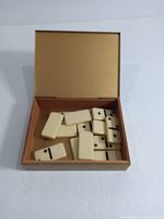 Open vintage tin box showing ivory domino tiles inside, with one tile in front showing a single black dot.