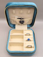 Open velvet jewelry case showing three silver rings and silver pendant with blue sapphires on cord inside compartments