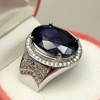 Close up of ring showing large oval blue sapphire, surrounded by halo of small clear stones, set in silver band with additional small stones on shoulders.