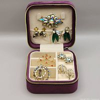 Top-down view of the velvet jewelry case open showing four pairs of earrings on top flap and brooches and pendant with chain in compartments below.