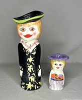 Front view of two ceramic vases designed as female figures, one tall in a black dress and hat, one small with colorful clothes and purple hat