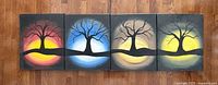Four paintings arranged in a row, showing four leafless black trees on colored gradient backgrounds ranging from yellow, blue, beige, and yellow again. All paintings are unframed canvas stretchers.