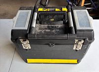 Front angled view of the black Stanley toolbox showing metal latches, yellow base, central handle, and translucent plastic compartments on top.