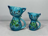 Pair of turquoise vintage ceramic cat-shaped measuring cups with floral decorations, different sizes showing the front side.