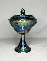 Front view of vintage Indiana Glass blue iridescent glass lidded jar on pedestal base featuring grape and leaf relief patterns.
