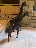Two dark brown metal giraffe figurines on carpeted floor near fireplace with brick background, showing size difference and finish.