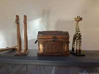Wide shot showing two hand carved wood giraffes on a wooden base, vintage wicker chest with metal accents and clasp, and yellow and black painted wooden giraffe figurine on separate base.