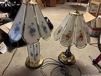 Two vintage Tiffany style glass and brass table lamps placed on floor in basement. One lamp has 6 panels with blue flowers, the other has 5 panels with pink and orange roses.