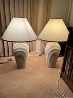 Pair of vintage cream ceramic table lamps with ribbed texture bases and cream fabric shades illuminated and turned on, placed on carpeted floor.