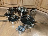 Full set of 6 stainless steel pots and pans with glass lids, alongside a stainless steel grater and potato ricer, all displayed on a wooden surface.