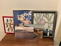 Five art pieces, three larger frames behind, two smaller photographic plaques in front, placed on wooden surface