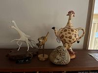 Display on wooden surface showing the four main items: white resin giraffes sculpture on a wooden base, burl wood carved crane, painted rock with giraffes, and whimsical giraffe teapot with lid.