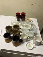 Top-down photo showing all 20 items arranged on a white table, including red and clear glass tumblers and various vintage ceramic mugs
