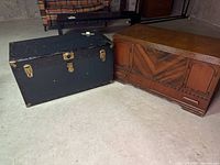 Both the wooden cedar chest and metal blue trunk side-by-side on the ground in a basement area.