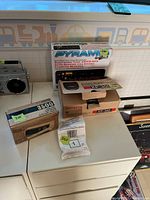 Four boxed items on white surface: Pyramid car stereo cassette player box, Aiwa XP-300 CD player box, electric Digi clock box, and sealed American Standard pressure balance spool package.