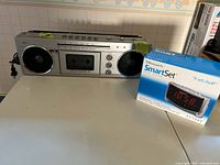 Candle FM/AM radio with cassette player and Research Smart Set dual alarm clock in original box on white countertop.
