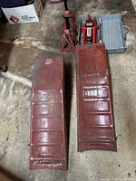 Overall view of two red metal vehicle ramps, hydraulic trolley jack, and two axle stands on floor.