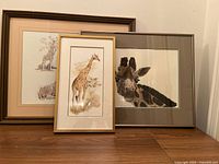 Three framed giraffe art pieces displayed on floor, showing overall style, color, and framing
