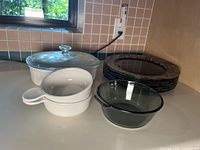 Photo of Corning Ware casserole dish with glass lid, two white Corning Ware soup bowls, clear Pyrex bowl, and stacks of Arcoroc dinner plates.