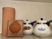 Natural clay wine cooler and bun warmer with sun motif, and two Corningware coffee pots with different decorative motifs on a shelf.