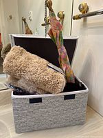 Photo of a textured fabric storage box with a body roller reclining on top, and a colorful umbrella inside the box, in a bathroom setting with a bathtub and towel bar visible