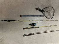 Full view of three fishing rods laid out on the floor with attached reel and fishing net placed next to them.
