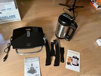 Photo showing the George Foreman Grill, SmartLiving Soup Maker, three black utensils, power cords, and recipe booklets on a wooden floor.