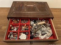 Top view of wooden jewelry box showing compartments and mixed men’s jewelry items including cufflinks, tie clips, and watch parts inside.