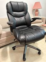Full view of a black leather swivel office chair against a beige couch and pink lamp background.